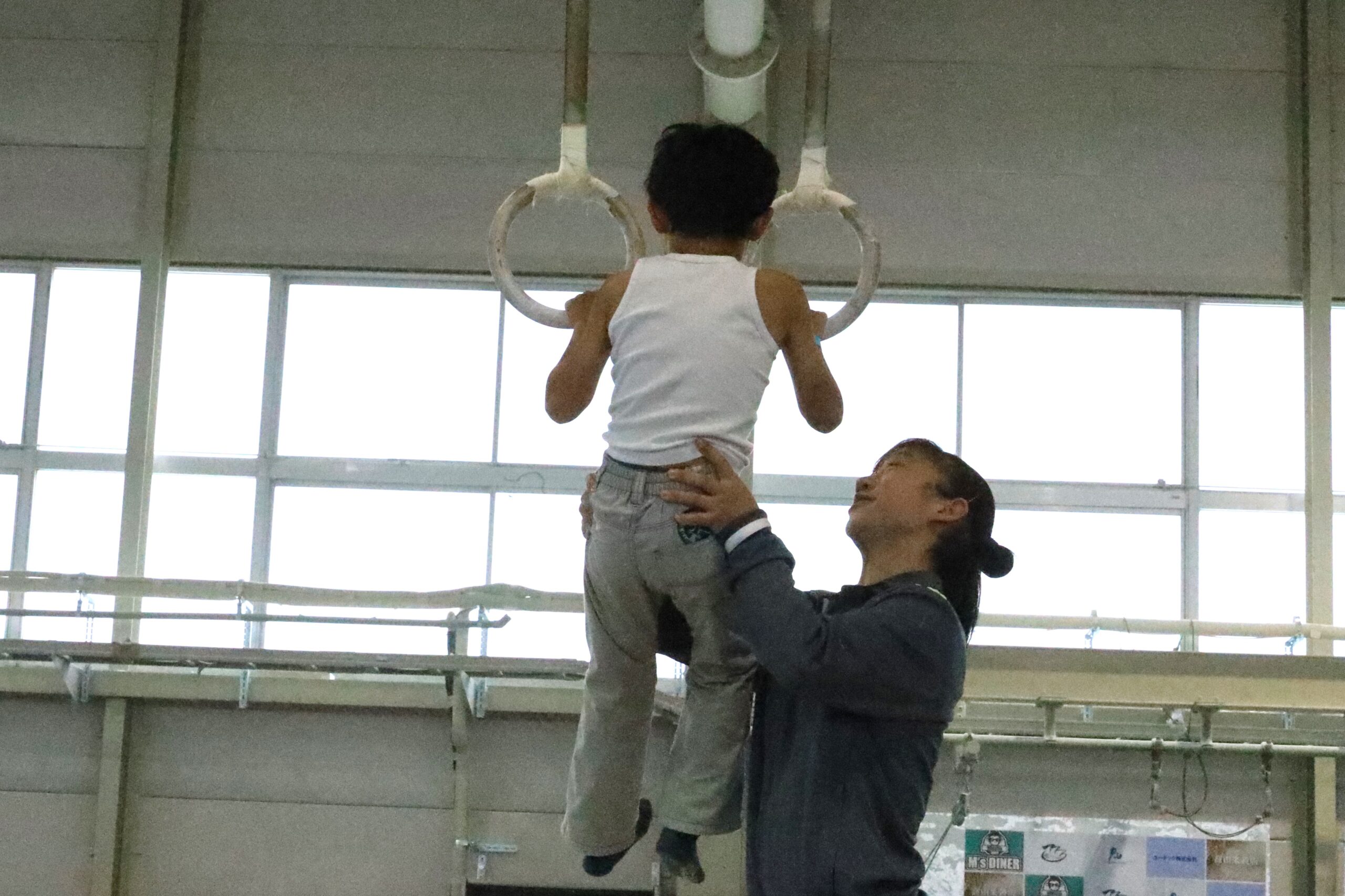 体操競技部員に抱きかかえられ、つり輪にぶら下がる男児